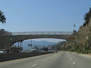 Several houses line the west side of California 1, and the Palisades line the east side as a pedestrian bridge crosses the highway. From here, California 1 proceeds northwest along the coast toward Malibu.