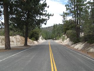 California 2 (Angeles Crest Highway) proceeds east through conifer forests and high-altitude vegetation. The highway has some long stretches of relatively straight and flat pavement, while other highway segments wind and curve through switchbacks and sharp turns. This view is taken near the Devils Canyon trailhead, about midway between Angeles Forest Highway N-3 and California 39.