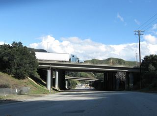 Historic Sierra Highway (old U.S. 6) proceeds southwest under California 14 (Antelope Valley Freeway), then curves southwest to meet Foothill Boulevard , then pass under the Interstate 5 (Golden State Freeway) Truck Bypass (but not the main lanes). At the intersection with The Old Road, U.S. 6 and U.S. 99 will turn south onto San Fernando Road en route to Sylmar and San Fernando. As this section of Sierra Highway was built originally as part of the Ridge Route, which later became and was replaced by U.S. 99, this section of U.S. 6 is quite old. The bridge at Tunnel Station was built in 1911 and widened in 1936; it spans over a railroad. Turn right here for U.S. 99/Newhall Alternate (built 1930, now called The Old Road) north toward Saugus or left for San Fernando Road (old U.S. 6-99) south to Los Angeles. Above, the massive Interstate 5 and California 14 interchange takes up most of this narrow valley. Trucks and cars scream or creep by depending on the severity of traffic congestion. San Fernando Road (old U.S. 6-99) is situated between the main lanes and truck bypass of Interstate 5 until it passes under the main lanes south of here.