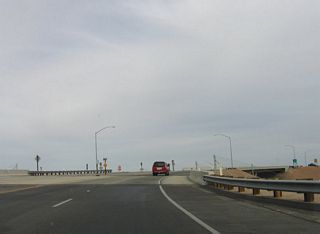 Now crossing a drainage ditch, California 7 north prepares to cross over and then merge with the two northbound lanes of commercial traffic coming from the commercial port of entry. The northbound route will turn north to join the trucks via a loop ramp (ahead).