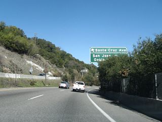 After the frontage road intersection, the California 17 freeway resumes again. This sign, which is mostly obscured by foliage, is located near the gore point for Exit 19, Santa Cruz Avenue north. Through traffic to San Jose should use the right two lanes.