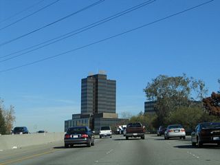 The high rises of the PruneYard come into view on northbound California 17. The PruneYard is a sprawling shopping center and business park built in 1964 located in eastern Campbell at the intersection of East Campbell Avenue and Bascom Avenue. The high rises are distinctive since they are the tallest buildings in this area outside of San Jose. When they were built, the PruneYard towers were the tallest buildings between Los Angeles and San Francisco. Today, a wide variety of stores are located here, as well as plenty of office tenants. Use Exit 25/Hamilton Avenue east to Bascom Avenue south to the PruneYard. The installation of a fourth lane (auxiliary/exit only) completed between 2004 and 2008 necessitated the replacement of the sign.