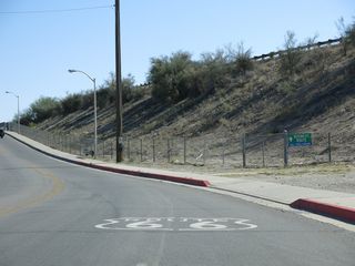 Affixed to a chain link fence, a business route sign appears along Broadway Street  south by the pedestrian tunnel below I-40 west to California Avenue.
