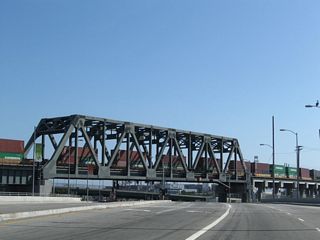 A large truss bridge takes the Alameda Corridor railroad line above both Dominguez Channel and the intersection joining Henry Ford Avenue with Pier A Way.