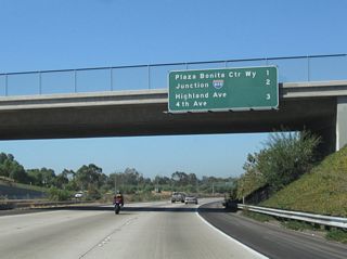 Passing under Woodman Street, this mileage sign along California 54 west provides the distance to the next three exits: Exit 3, Plaza Bonita Center Way and Reo Drive; Exit 2, Interstate 805 (Jacob Dekema [Inland] Freeway); and Exit 1D, Highland Avenue north and Fourth Avenue south.