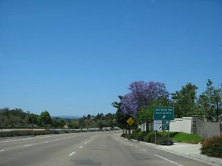 A blooming jacaranda tree serves as the backdrop for the advance sign prior to the off-ramp to Exit 9C, Rancho Carmel Drive north to the Carmel Mountain Ranch community of San Diego and Sabre Springs Parkway south to the Sabre Springs community of San Diego.