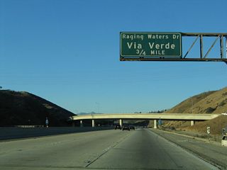 Before leaving the Kellogg Interchange, this sign identifies the next exit from California 57 (Orange Freeway) north as Exit 22, Via Verde and Raging Waters, 0.75 mile. This is the first exit to serve the city of San Dimas. Ahead, traffic from Interstate 10 west merges onto California 57 north. This ramp was closed for a time on April 25, 2010, after a landslide brought rocks and mud onto the transition ramp. This ramp used to bring traffic from Interstate 10 west to Interstate 210 west before 210 was rerouted.1