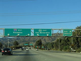 Northbound California 57 (Orange Freeway) reaches Exit 25A, Auto Centre Drive. As for through traffic, the left lanes default onto westbound Interstate 210, while the right lanes head east along California 210. Wires can partially obscure the legibility of the sign.