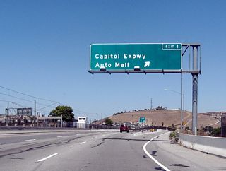 Northbound California 87 (Guadalupe Parkway) reaches Exit 1, Santa Clara County Route G-21 (Capitol Expressway). A transit station for the light rail (which follows the California 87 median from here north to Interstate 280) can be seen in the middle of the freeway.