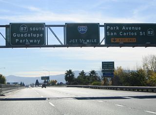 The right two lanes separate at Exit 6 A to Park Avenue and California 82 (West San Carlos Street) and Exit 5 to Interstate 280 (Sinclair Freeway). Exit 6 A connects to Downtown San Jose. Park Avenue crosses Guadalupe River Park ahead of Plaza de Cesar Chavez. The Paseo de San Antonio Walk continues east past this point as a pedestrian plaza to San Jose State University.