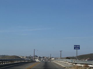 A curving bridge carries First Avenue over Riverside Drive and the Mojave River, which remains apparently dry most of the time. Old bridge railing is obscured by newer standard guardrail. At the end of the bridge is the railroad depot, which includes a Route 66 Museum.