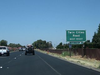 In typical style, the county designation and street name are only mentioned once, on this auxiliary sign on the approach to Exit 277. CR E-13 is an east-west county route that begins here at California 99 and extends west along Twin Cities Road and River Road to Walnut Grove, ending at California 160 at the west end of the Walnut Grove Bridge. The portion of CR E-13 that follows River Road is situated mostly on a levee overlooking the Sacramento River in the California Delta region.