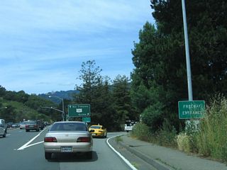A freeway entrance shield assembly is partially obscured by grass as westbound California 131 comes to an end. Straight ahead, Tiburon Boulevard becomes Blithedale Avenue, which serves the city of Mill Valley.