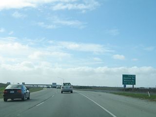 Traffic from westbound California 156 merges onto California 1 (Cabrillo Highway) south. Another sign for upcoming Exit 414 (to Nashua Road and Molera Road) appears in the distance. The left two lanes will merge with the incoming California 1 lanes, and the freeway will travel the remaining distance along the coast to Monterey and Pacific Grove.