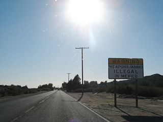 The typical warning signs for traffic entering Mexico appear along California 186 as it continues southwest toward Andrade and Mexico. The first sign advises of the permit required to export firearms, and this sign warns that guns and ammunition are illegal in Mexico. After these signs, slow down. Parked cars will appear on both sides of the highway.