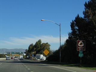 Another California 237 east shield assembly is posted along with a historic Juan Bautista de Anza route shield after the on-ramp from Middlefield Road and Maude Avenue. The U.S. 101 interchange can be seen in the distance.
