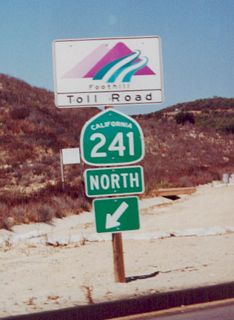 The Orange County Toll Roads generally use a different style of freeway entrance signage than most California freeways. This particular freeway entrance assembly, located along Oso Parkway (Exit 3), uses the standard Foothill Corridor logo in lieu of a freeway entrance sign. Also notice the slightly angular look to this California state shield. At the time this photo was taken, this was the first exit along northbound California 241; since then, the toll road has been extended south to Antonio Parkway.  A further extension of the toll road to Interstate 5 near the Basilone Road exit in San Diego County is proposed, but it has encountered environmental and military opposition.