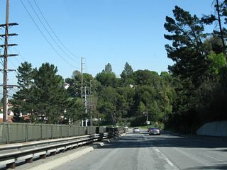The roadway curves northeast and continues downhill. A concrete median barrier is placed between the two directions of traffic to prevent crossover accidents.