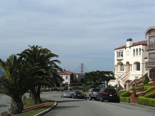 Palms dominate the median as the Golden Gate Bridge comes into view along El Camino del Mar in Sea Cliff. The road turns right at the stop sign ahead.