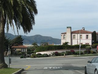 Palms dominate the median as the Golden Gate Bridge comes into view along El Camino del Mar in Sea Cliff. The road turns right at the stop sign ahead.