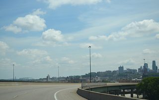 Curving east toward the Kansas River, I-670 spans the I-70 interchange with Pacific Avenue and several railroad lines along a pair of three lane viaducts. Visible along the northeastern horizon is the Broadway Bridge for U.S. 169.