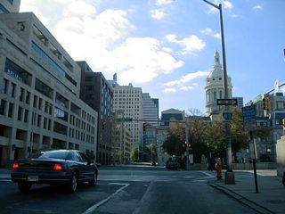 An older Interstate 83 trailblazer, since replaced, directs Fayette Street westbound drivers onto Gay Street northbound for the Jones Falls Expressway northbound on-ramp at the Fallsway. The rotunda of Baltimore City Hall lies to adjacent to the Fayette and Gay Street intersection.