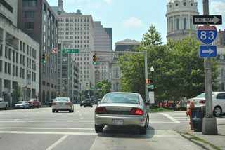 An older Interstate 83 trailblazer, since replaced, directs Fayette Street westbound drivers onto Gay Street northbound for the Jones Falls Expressway northbound on-ramp at the Fallsway. The rotunda of Baltimore City Hall lies to adjacent to the Fayette and Gay Street intersection.
