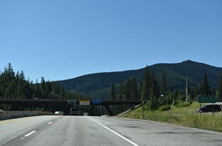 Interstate 90 enters Mineral County, Montana and Mountain Time Zone at Lookout Pass (elevation 4,725 feet). Roland Point (elevation 5,374 feet) in Shoshone County, Idaho rises beyond the Mineral County Route 174 overpass.