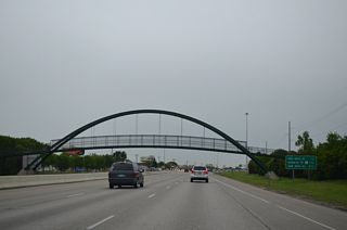 A steel arch pedestrian bridge spans I-94/U.S. 52 between the adjacent Brunsdale and Lewis & Clark neighborhoods west of University Drive.