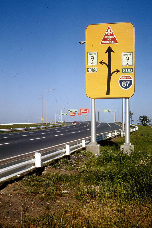 La Prairie.  <br><br>

<i>These are still the old Quebec shields, before the grand renumbering.  What is now Autoroute 10, was still the Autoroute des Cantons de L’est, and its shield was a red triangle.  What really fascinated me was that the guide signs specific to that route in the distance were also red.  I know that other toll autoroutes had a similar triangular shield in different colors, including blue and green (BGS, BBS and BRS – sheesh!).  Would those guide signs be matched to the toll road color?  Of course an Interstate shield in Canada is always something special.</i>
