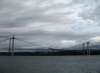 Looking at Tacoma Narrows Bridge during construction of the eastbound span from the view area off Lucille Parkway NW. Sections of the eastbound Tacoma Narrows Bridge deck awaited placement on a ship near the north tower.
