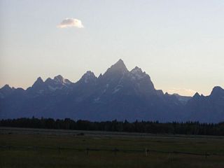 At Moran Junction, the majestic Grand Tetons dominate the skyline, with its jagged peaks backlit by a setting sun in the early evening.